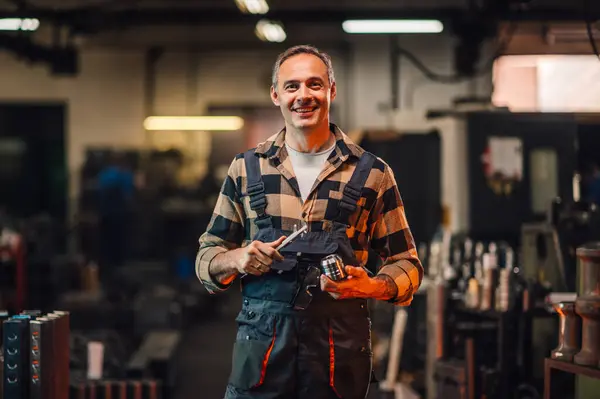 Metalurji teknisyeninin elinde bir alet ve silindir makine elementi olan kameraya gülümseyen bir resmi. Çeşitli metal şekilli ve makineli imalat tesisinin bulanık arka planı.