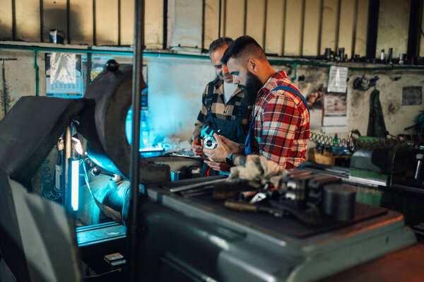 Two metallurgists examining a metal fitting prepared on a lathe machine. A senior and a young worker with a beard, wearing overalls and plaid shirts. Factory, manufacture setting.