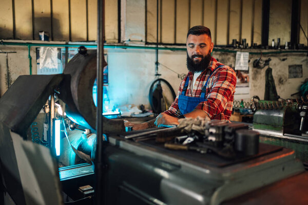 Young male metallurgist with a beard working on a milling machine to create a new metal part. He wears a blue worker uniform and a plaid shirt. Old workshop, manufacture, artisan, craftsman.