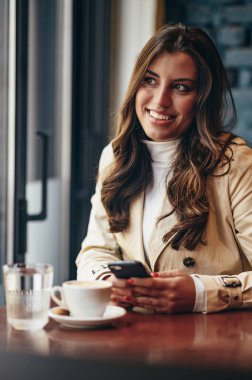 Pencerenin yanındaki bir kafede otururken akıllı telefon kullanan güzel bir genç kadın.