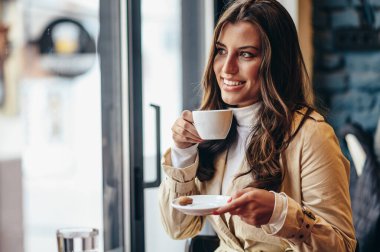 Güzel bir genç kadın pencerenin yanındaki bir kafede otururken akıllı telefon kullanıyor ve kahve içiyor.