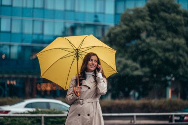Güzel bir genç kadın, şehirde şiddetli bir yağmur sırasında akıllı telefon kullanıyor ve elinde sarı bir şemsiye tutuyor.