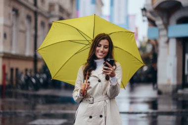 Güzel bir genç kadın yağmurlu bir günde şehirde sarı bir şemsiye tutuyor ve akıllı telefon kullanıyor.