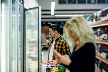 Genç mutlu çift markette akıllı telefon kullanıyor alışveriş arabalarıyla market alışverişi yaparken ürün seçiyorlar.