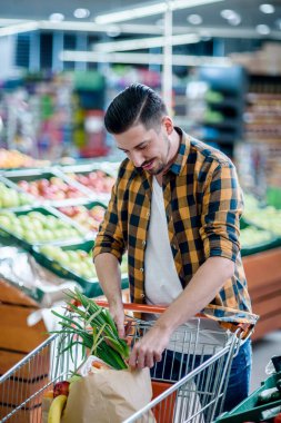 Süpermarkette genç yakışıklı bir adam market alışverişi yaparken ürün seçiyor ve alışveriş arabası tutuyor.
