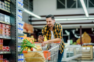 Süpermarkette ürün seçen ve elinde alışveriş arabası ve market alışverişi tutarken akıllı telefon kullanan genç yakışıklı bir adam.