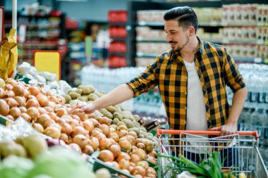 Süpermarkette genç yakışıklı bir adam market alışverişi yaparken ürün seçiyor ve alışveriş arabası tutuyor.