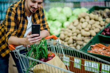 Süpermarkette akıllı telefon kullanan, ürünleri seçen ve alışveriş arabasıyla alışveriş yapan genç yakışıklı bir adam.