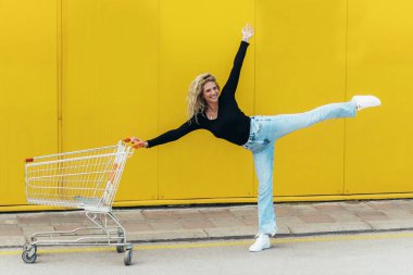 Süpermarket arabası olan genç ve güzel bir kadın bale pozisyonunda sarı arka planda duruyor.