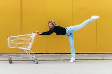 Süpermarket arabası olan genç ve güzel bir kadın bale pozisyonunda sarı arka planda duruyor.