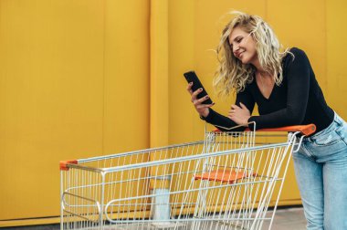 Güzel, genç bir kadın alışveriş arabasının arkasındayken akıllı telefon kullanıyor.