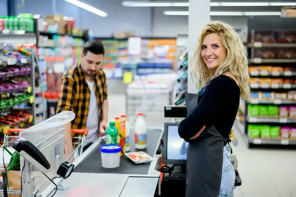 Güzel genç kasiyer süpermarkette çalışırken gülümsüyor ve önlük giyiyor.