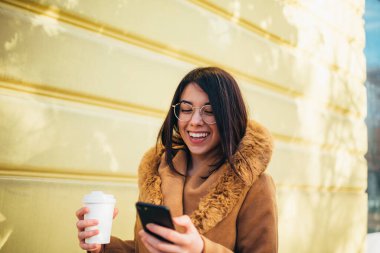 Genç İspanyol kadın akıllı telefon kullanıyor ve dışarıda yürürken kahve içiyor.