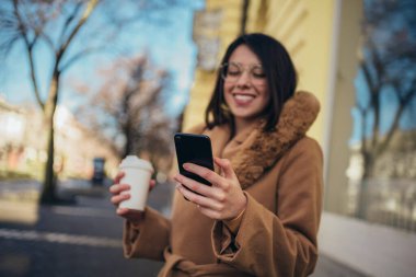 Genç İspanyol kadın şehirde yürürken akıllı telefon ve kahve servisi kullanıyor.