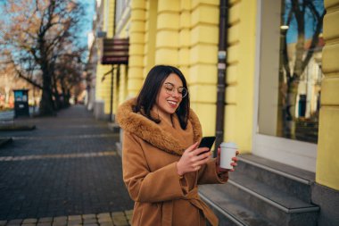 Genç İspanyol kadın şehirde palto giyerken akıllı telefon kullanıyor.