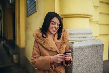 Genç İspanyol kadın şehirde palto giyerken akıllı telefon kullanıyor.