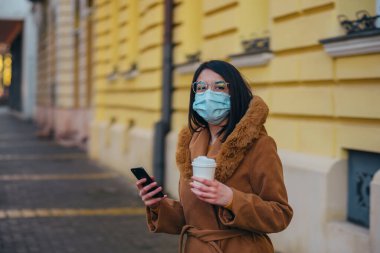 Akıllı telefon kullanırken ve dışarıdayken kahve içerken koruyucu maske takan genç İspanyol kadın.
