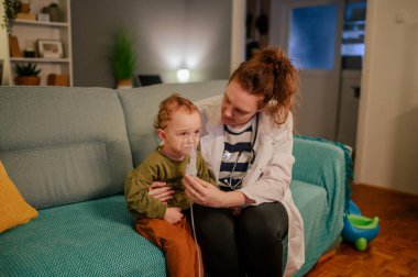 Bir doktor, solunum hastalığı olan küçük hasta hastasıyla kanepede oturuyor ve onu soluyor. Bir çocuk doktoru bronşiti olan hasta bir çocuğa tıbbi yardım ve destek veriyor. Solunum.