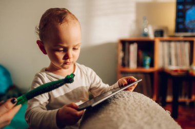 Odaklanmış küçük bir çocuk çizim tahtasını tutuyor ve evde ailesiyle kalırken üzerine bir şeyler karalamaya hazırlanıyor. Küçük bir çocuk elinde bir çizim tahtası tutuyor ve çizmeye hazırlanıyor..
