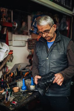 Yaşlı bir zanaatkar, ayakkabıcı atölyesinde elinde botlarıyla dikilip onları dikizliyordu. Küçük bir işletme sahibinin portresi. Ayakkabıcı atölyesinde duruyor ve botlarını inceliyor..