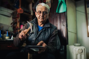 Eski bir kunduracı atölyesinde oturuyor ve ayakkabı tabanını tamir etmek için çekiç kullanıyor. Küçük atölyesinde ayakkabı parçalarıyla çalışan usta bir zanaatkarın portresi. Sanatçılık konsepti. Boşluğu kopyala.