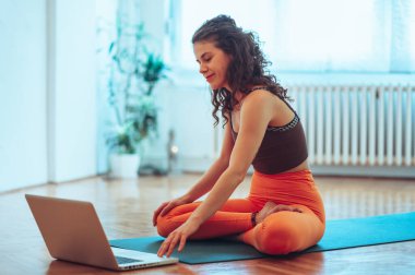 Nilüfer çiçekli bir poz veren genç bir kızın parlak bir stüdyoda yoga minderinde dizüstü bilgisayarında bir ders hazırlarken yan görüntüsü. Yoga stüdyosu ve bitkilerin bulanık arkaplanı. Boşluğu kopyala.