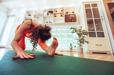 Parlak bir stüdyoda başını dizine yaslarken yoga minderinin üzerinde esneyen siyah saçlı kıvırcık bir kızın düşük açılı görüntüsü. Bir bacağını öne uzatıyor ve ayağını esnetiyor. Boşluğu kopyala.