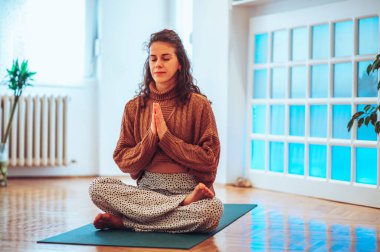 Kafkas saçlı güzel bir kızın lotus pozunda yoga minderinde otururken ve elleri dua pozunda göğsünün önünde otururken geniş bir kare. Gözleri kapalı ve yüzünde nazik bir gülümseme.