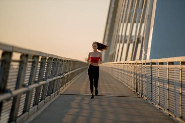 Gün batımında köprüde koşan çekici genç bir kadının fotoğrafı.