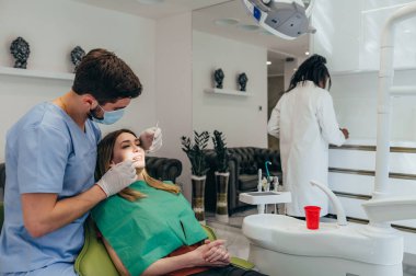 Erkek dişçi, asistanının yardımıyla dişçi koltuğunda oturan bir kadının dişlerini düzeltiyor. Diş hekimi hasta dişlerini muayene ediyor ve diş hekiminde diş muayenesi yaptırıyor..