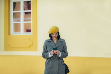 Akıllı telefon kullanan, sarı bereli ve gri montlu güzel bir kadın şehrin sokaklarında yürüyor.