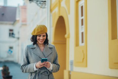 Çekici bir genç kadının gri ceketli ve sarı bereli bir şehri keşfederken çekilmiş.