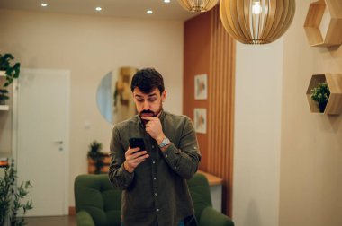Thoughtful caucasian man holding smartphone and typing a message, chatting online, using dating app while standing in the living room at home. Young joyful man with beard using mobile phone. clipart