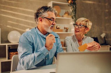 Üst düzey bir çift evdeki masada dizüstü bilgisayar ve kredi kartıyla alışveriş yapıyor. İnternet sitesi ve banka kartıyla satın alma, otel rezervasyonu. Olgun çift çevrimiçi ödeme sistemini öğreniyor.