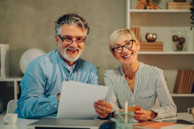 Yetişkin evli bir çiftin portresi fatura ya da mali durumları kontrol ediyor ve evdeki masada otururken dokümanları inceliyor. Ev giderlerini hesaplayan yaşlı eşler. Kameraya bakıyorum..