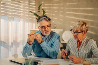 Üst düzey çift evdeki masada birlikte otururken gazeteleri kontrol ediyor. Gülümseyen olgun eşler, internetten ödeme yapmadan önce faturaları ödemeye başlıyorlar. Kredi kartı tutan bir adam. Boşluğu kopyala.