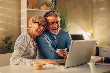 Evde dizüstü bilgisayarla oturmuş gülen ve kaynaşan mutlu bir çift. Gülümseyen kıdemli eşler internette gezinirken sarılıp eğleniyorlar ve birlikte vakit geçiriyorlar..