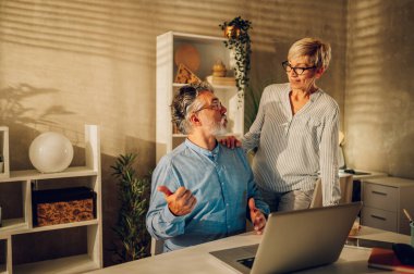 Evde dizüstü bilgisayar kullanırken birbirlerine bakıp gülen olgun bir çiftin portresi. Gülümseyen kıdemli eşler internette gezinirken ve birlikte vakit geçirirken eğleniyorlar. Boşluğu kopyala.
