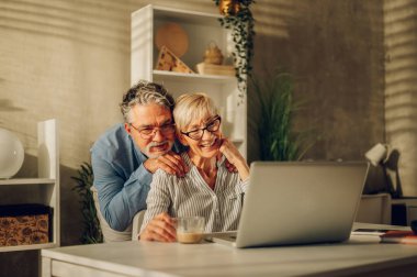 Evde dizüstü bilgisayar kullanırken gülüp kaynaşan mutlu bir çiftin portresi. Gülümseyen yaşlı karı koca internette gezinirken eğleniyor ve sarılıyorlar ya da iyi haberler okuyorlar. Boşluğu kopyala.