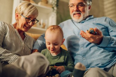 Büyük anne ve büyük baba, çocuk torunla oynuyorlar ve evdeki kanepede otururken birlikte vakit geçiriyorlar. Mutlu olgun eşler birlikte torun bakıcılığı yapıyor. Bir çocuğa odaklan..
