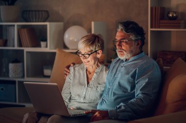 Evdeki kanepede oturan ve birlikte dizüstü bilgisayar kullanan yaşlı evli bir çift. Gülümseyen olgun koca ve karısı internet üzerinden alışveriş yaparken ya da çalışırken eğleniyorlar..
