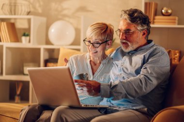 Bir kanepede dizüstü bilgisayarla oturmuş gülüp kaynaşan olgun bir çiftin portresi. Gülümseyen kıdemli eşler internette gezinirken ve evde birlikte vakit geçirirken eğleniyorlar. Boşluğu kopyala.