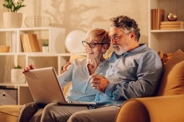 Mutlu, olgun bir çift evde kanepede oturup dizüstü bilgisayar kullanırken gülüyor. İnternette gezinirken ve birlikte vakit geçirirken gülümseyen yaşlı eşlerin portresi. Boşluğu kopyala.