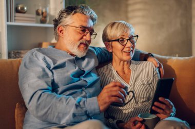 Mutlu son sınıf evli çift evde dinlenirken akıllı telefon kullanıyor. Gülümseyen eşler evde cep telefonlarıyla eğleniyorlar ve internet veya online alışveriş yapıyorlar..