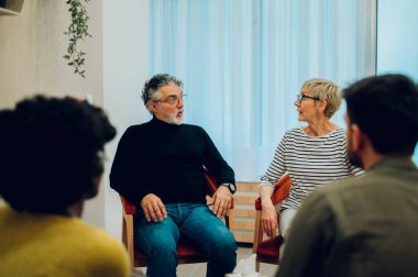 Ruh sağlığı merkezindeki bir toplantıda kıdemli bir kadınla konuşan kıdemli bir adam. Grup terapisi katılımcıları bir çemberin içinde oturup konuşuyorlar. Yaşlılar zihinsel sağlıklarına dikkat ediyorlar.