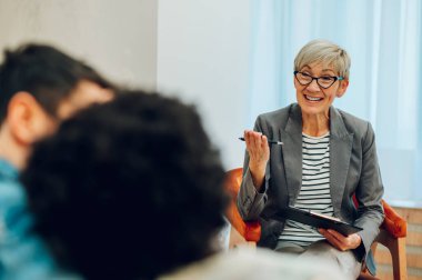 Gülümseyen olgun bir kadın doktorun portresi, ofisinde not alırken çeşitli müşterilerinin zihinsel ve evlilik problemlerini dinliyor. Psikoterapi danışmanlığı.