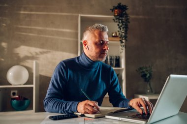 Orta yaşlı bir adam modern işyerindeki bilgisayarında çalışıyor ve not alıyor. Gri sakallı ve saçlı bir iş adamı. Ev ofisinin modern bir çalışma yerinde çalışıyor..