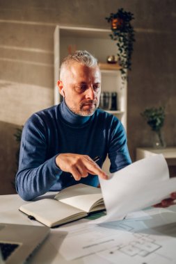 Keyifli orta yaşlı beyaz adam bir ofis masasında otururken sözleşmeleri ve iş belgelerini okuyor. Evde çalışan iş adamları kavramı. Balıkçı yaka kazak giyiyor. Boşluğu kopyala.
