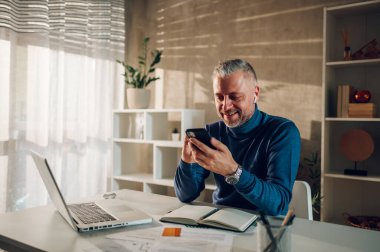 Ev ofisteki bir masada otururken uzaktan kumandayla ve akıllı telefonla çalışan orta yaşlı yakışıklı bir serbest çalışan. İş ve serbest çalışma. Bir ileti yazılıyor veya bir istemciye e- posta yeniden oynatılıyor.