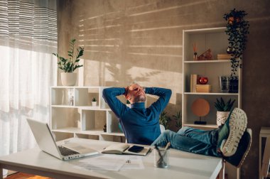 Neşeli, gri saçlı, orta yaşlı, beyaz bir adam ev ofisinde otururken masanın üstünde bacak tutuyor. Stresli bir işten sonra rahatlama kavramı. Boşluğu kopyala.